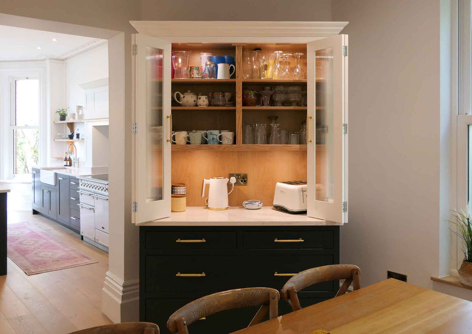 a handcrafted, bespoke shaker kitchen with a run of dark grey shaker cabinetry, a large shaker island, a freestanding white larder cupboard with double doors and oak spice racks and a freestanding dresser with a dark grey base and a white countertop cupboard with glazed bi-folding doors and oak shelving