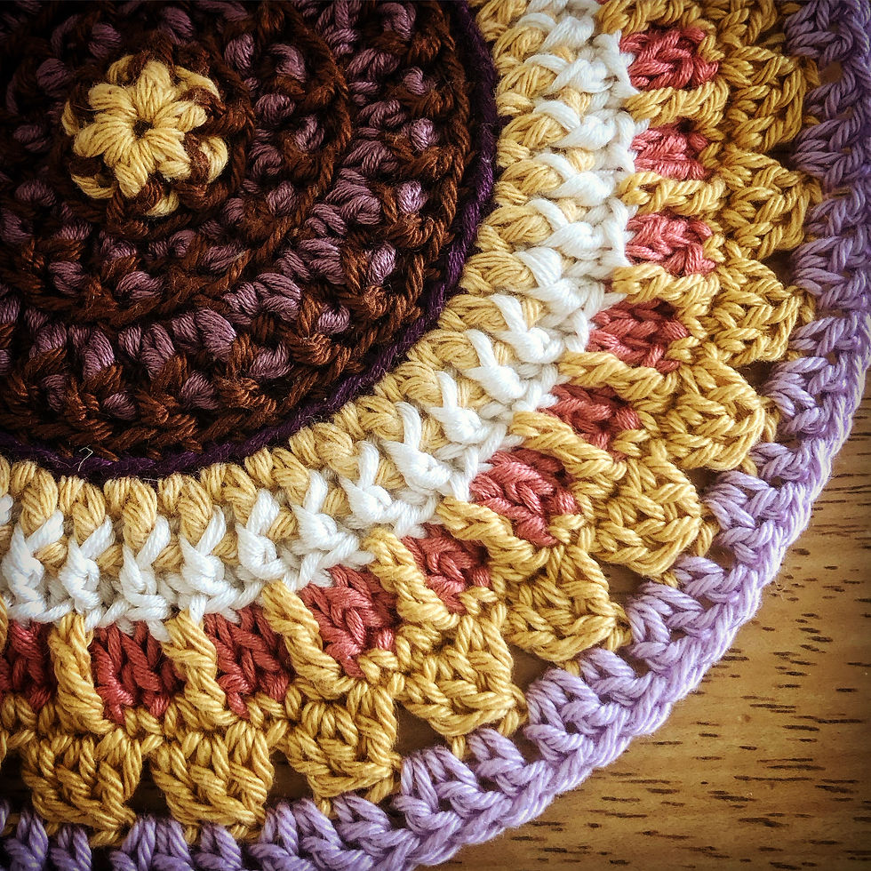 Crocheted mandala in brown, yellow, white, pink, and purple yarn, forming a circular, intricate pattern on a wooden surface.