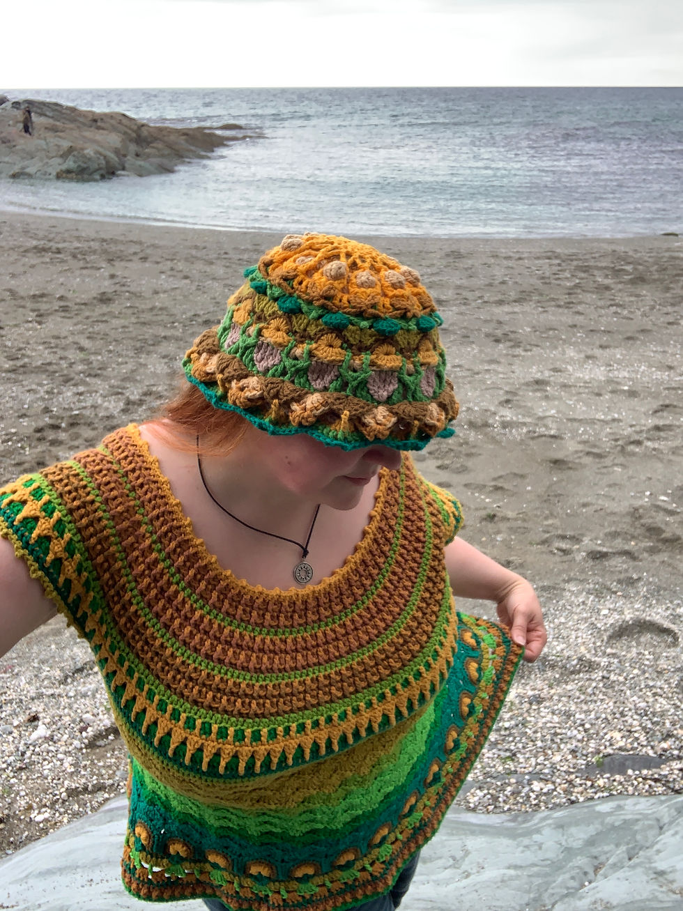 Person on a beach wearing a colourful crocheted hat and top, featuring green, brown, and yellow patterns. Sea and rocky shore in background.
