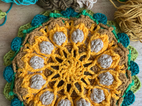 Colourful crochet mandala in yellow, green, brown on a wooden surface. Surrounded by skeins of yarn in green, beige, and mustard.