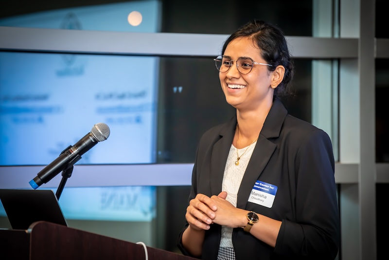 A businesswoman with long, dark hair, medium skin tone and glasses is presenting at a MIRTA event. She is wearing a black blazer and standing at a podium.