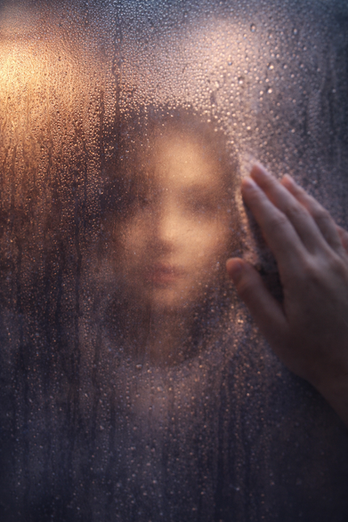 A hand clears condensation from a fogged window or mirror, revealing a soft, partially hidden face behind the glass.
