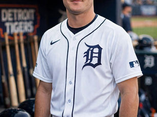 Smiling Detroit Tigers player Kevin McGonigle in white home uniform stands in the dugout, with bats, helmets, and Detroit Tigers text behind him.