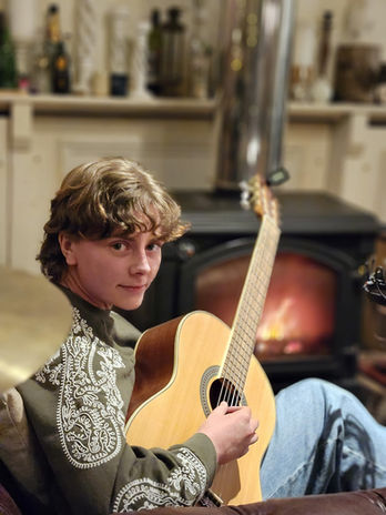 Luce sitting with an acoustic guitar in front of a glowing wood-burner, looking at the camera.
