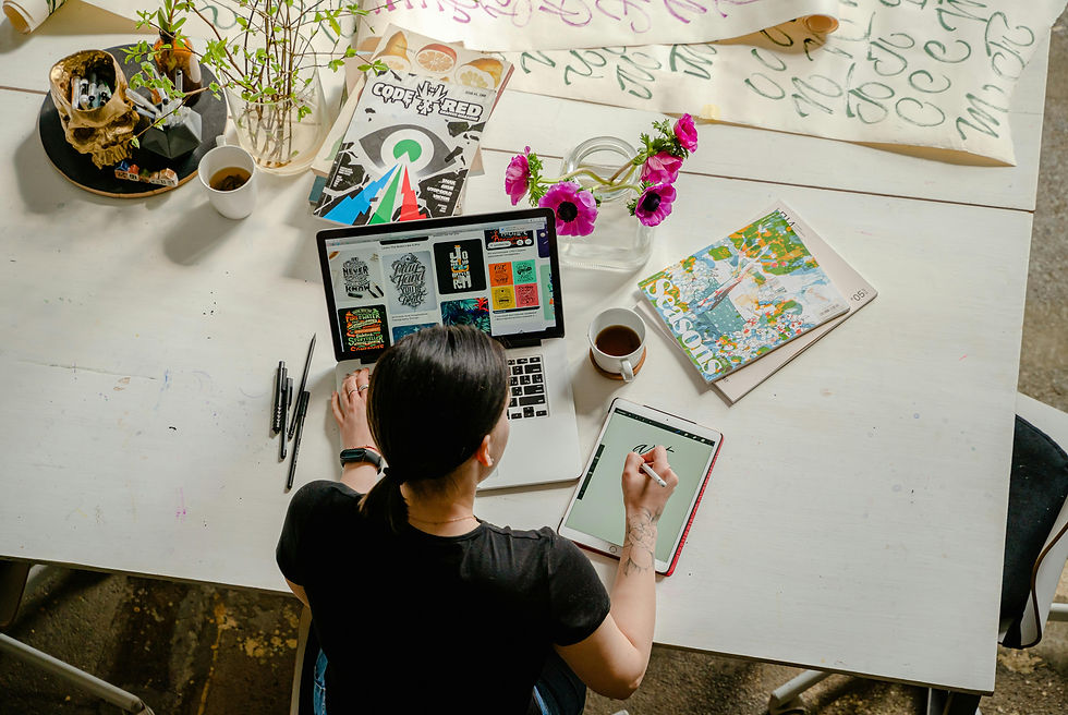 Person drawing on a tablet at a white table with art supplies, magazines, and flowers. Laptop screen shows graphic designs. Bright setting.
