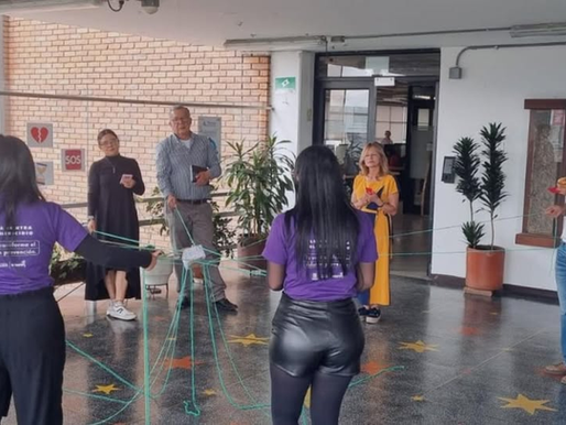 Un grupo de personas en un espacio interior semicubierto participa en una actividad comunitaria o taller. Varias personas, entre hombres y mujeres, forman un círculo mientras sostienen y entrelazan hilos verdes que crean una red en el centro. Dos mujeres en primer plano visten camisetas moradas; en la espalda de una de ellas se lee una inscripción relacionada con la lucha contra el feminicidio y la frase "Tu voz transforma el dolor en prevención". Al fondo se observan plantas decorativas, un piso con estrellas amarillas pintadas y una pared de ladrillo.
