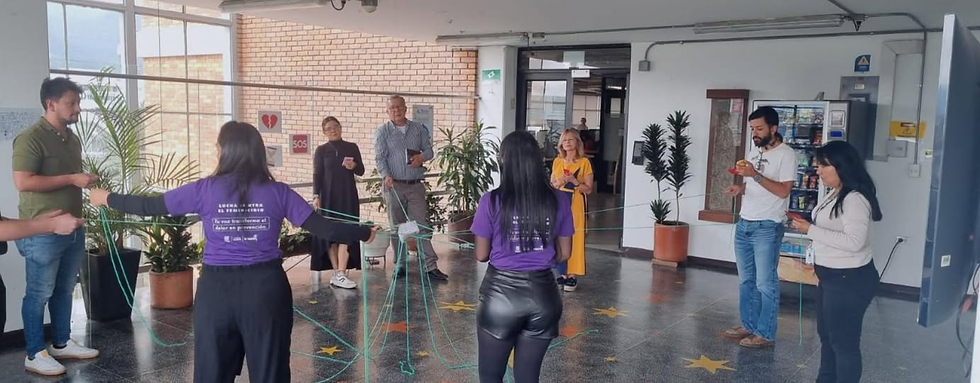 Un grupo de personas en un espacio interior semicubierto participa en una actividad comunitaria o taller. Varias personas, entre hombres y mujeres, forman un círculo mientras sostienen y entrelazan hilos verdes que crean una red en el centro. Dos mujeres en primer plano visten camisetas moradas; en la espalda de una de ellas se lee una inscripción relacionada con la lucha contra el feminicidio y la frase "Tu voz transforma el dolor en prevención". Al fondo se observan plantas decorativas, un piso con estrellas amarillas pintadas y una pared de ladrillo.