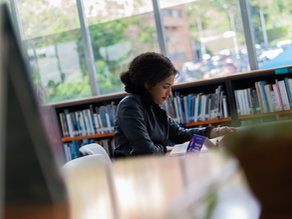 Mujer leyendo en una biblioteca rodeada de varias estanterías de libros y concentrada en su lectura