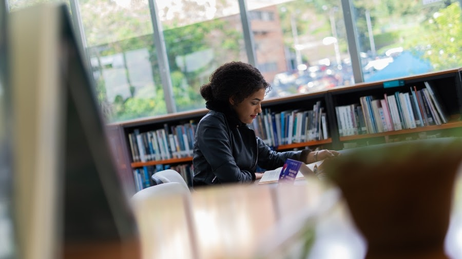 Mujer leyendo en una biblioteca rodeada de varias estanterías de libros y concentrada en su lectura