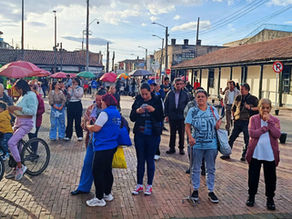 Multitud en plaza empedrada con bicicletas y sombrillas; gente espera junto a edificios coloniales bajo cielo soleado.