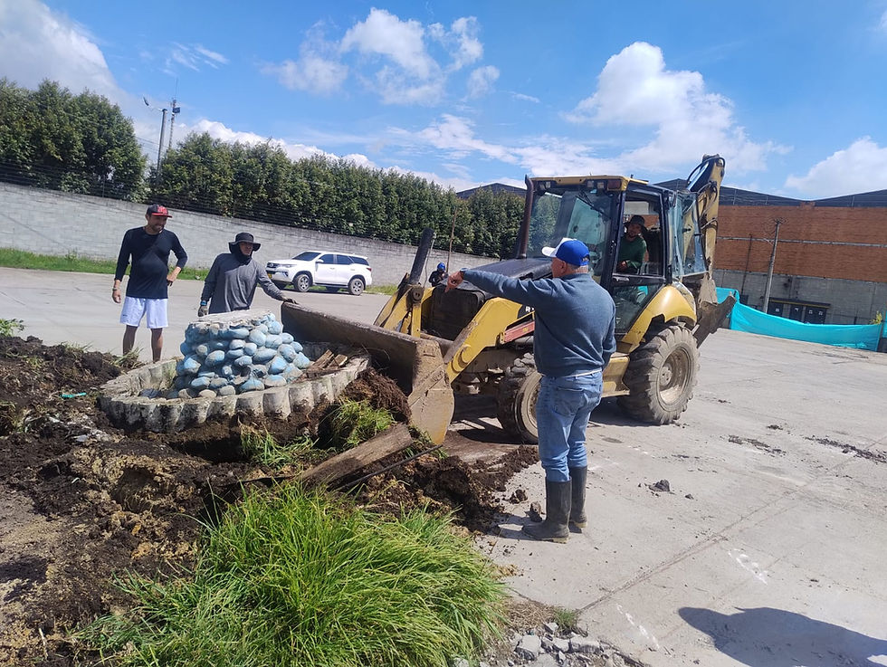 Operarios de construcción y maquinaria pesada trabajando en la remoción de una estructura de piedra decorativa en un estacionamiento. Un trabajador con gorra azul dirige a un conductor de retroexcavadora Caterpillar amarilla mientras otros supervisan la obra en un día despejado.