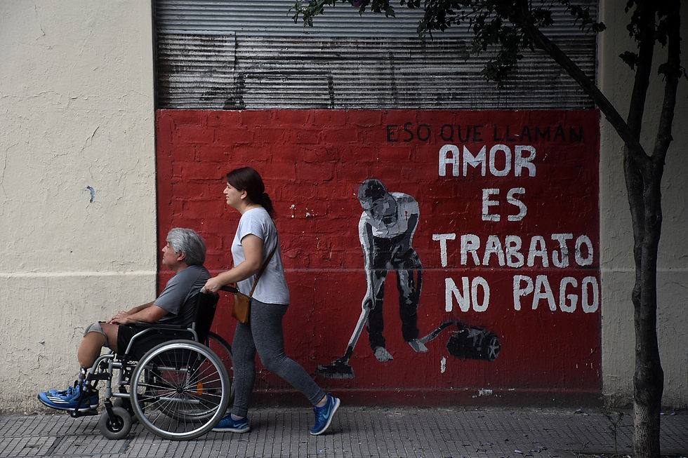 Mujer empuja a un hombre en silla de ruedas frente a un mural rojo que dice: Es lo que llaman amor, es trabajo no pago.