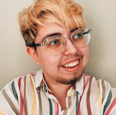 Photo of Sam Dylan Finch wearing glasses and looking up to his left, the viewer's right-hand side.
He's wearing a striped, collared shift with a white background and grey, green, red, orange, and yellow stripe.
He is smiling with facial hair and blonde hair on his head.