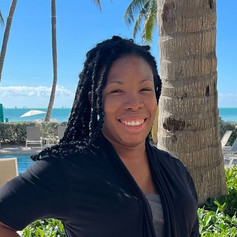 Fallon is looking at the camera, with her right hand on her hip smiling at the camera.
She's smiling with her long black hair draped over her black sweater and grey shirt.
In the background there is a palm tree, blue sky and ocean.