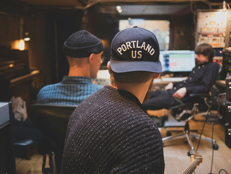 Three people in a recording studio. One wears a hat reading "PORTLAND US." Monitors and audio equipment visible. Casual, focused atmosphere.