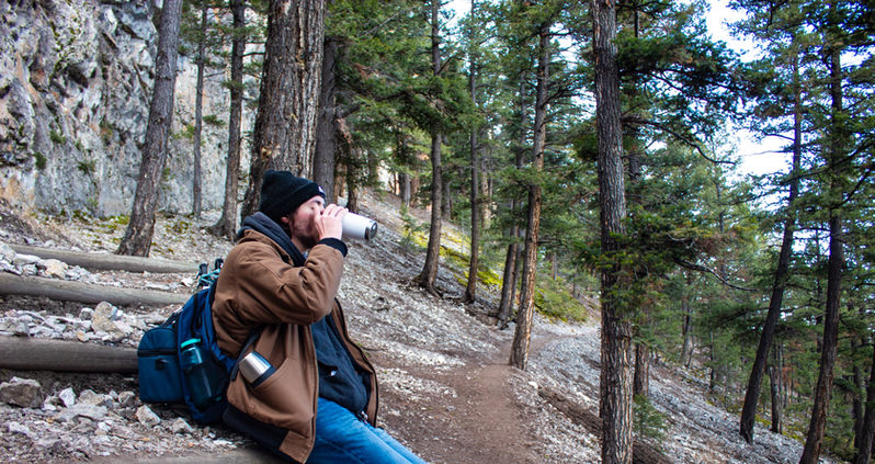 A hiker in a brown jacket and black beanie sitting on a wooden trail step, drinking from a silver mug.