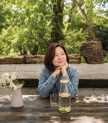 Loi Gesmundo, VA Mentor and founder of Loi Gesmundo Digital, seated outdoors at a wooden table with flowers and a water pitcher, resting her chin on her hands.