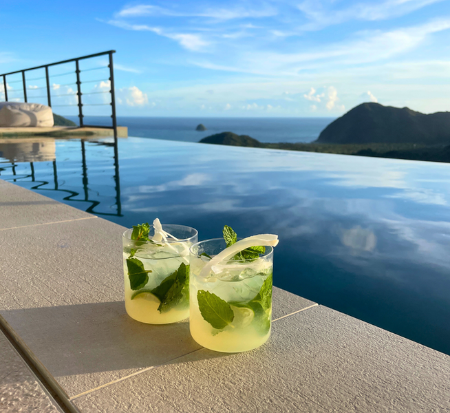 Zwei Cocktialgläser mit Kokos-Minz-Cooler gefüllt, stehen an einem Poolrand, Blick in eine Bucht.