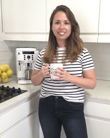 Woman in striped shirt holds a mug in a bright kitchen. Coffee machine and lemons on counter. She smiles, creating a cozy, inviting mood.