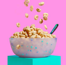 Cereal falling into a frosted bowl with a "Magic Spoon" labeled spoon, set against a bright pink background.