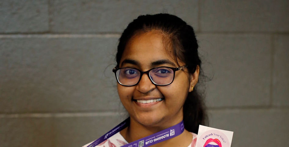 A young woman smiles as she holds up a pink “Unleash Your Voice” sticker and a purple DC Blogging Club lanyard.