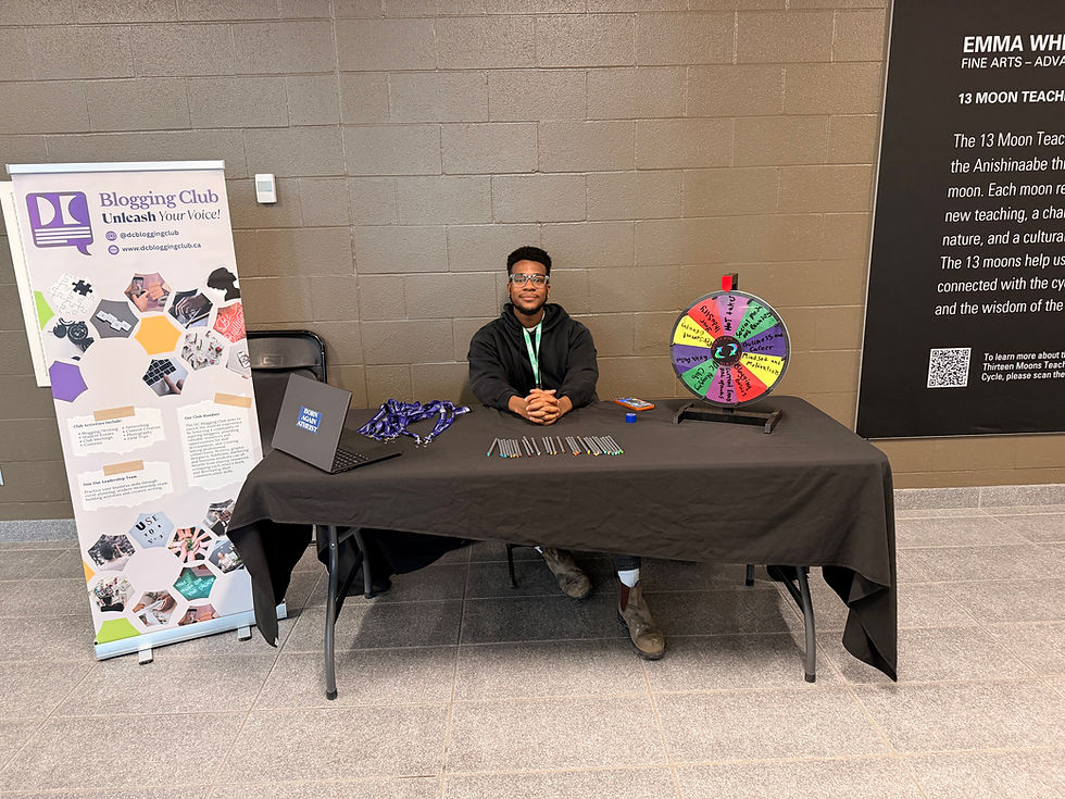 Jamal sits at a table with a colorful prize wheel, promoting the DC Blogging Club. A laptop, pens, and banner complete the setup. Brick wall backdrop.