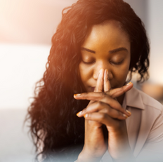 Woman with eyes closed, hands clasped in prayer or contemplation, indoors. Warm lighting, soft focus creates a calm and reflective mood.