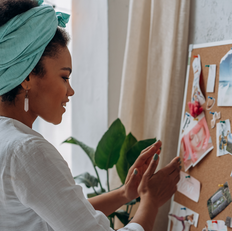 Woman in a white shirt and teal headwrap arranges images on a corkboard. Background shows a plant and curtain. Mood is focused.