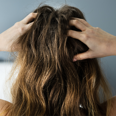 Person with long brown hair holds their head with both hands, set against a blurred gray and white background, conveying stress or frustration.