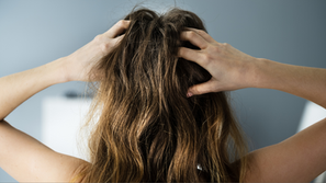 Person with long brown hair holds their head with both hands, set against a blurred gray and white background, conveying stress or frustration.