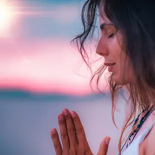 Woman in profile, eyes closed, hands in prayer. Serene expression. Sunset sky in pink and blue tones in the background.