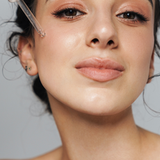 Woman with neutral expression applies serum to her cheek using a dropper. Close-up, clear skin, natural makeup, soft lighting.