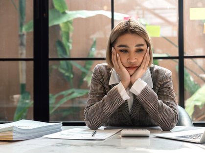 Woman in a plaid suit looks pensive, resting her head in hands at a desk with a laptop and papers. Large window with plants in background.