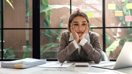 Woman in a plaid suit looks pensive, resting her head in hands at a desk with a laptop and papers. Large window with plants in background.