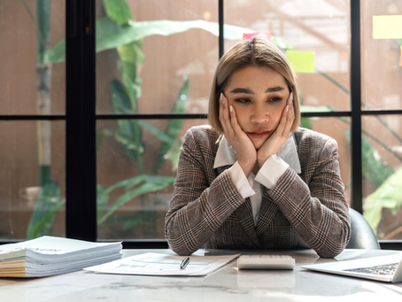 Woman in a plaid suit looks pensive, resting her head in hands at a desk with a laptop and papers. Large window with plants in background.