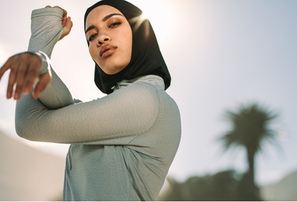 A woman in a gray top and black headscarf stretches outdoors. Sunlight creates a halo effect. Palm tree and clear sky in the background.