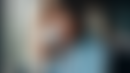 A person with curly hair sips from a white mug, wearing a blue shirt. They're seated in a cozy room with soft background lighting.