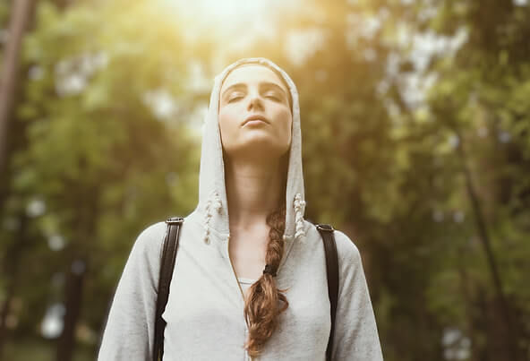 Woman in a gray hoodie with eyes closed stands peacefully in a sunlit forest. Backpack straps visible, serene mood.
