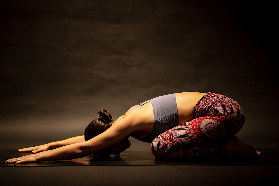 Woman in colorful leggings and a sports top doing child's pose on a mat in a dimly lit room, creating a calm and meditative mood.