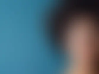 Woman with curly hair and glasses gazes thoughtfully to the side against a blue background, suggesting contemplation or curiosity.