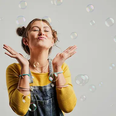 Woman in yellow sweater and denim overalls joyfully blowing bubbles against a gray background. Playful mood with colorful bubbles.
