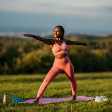 Person in pink workout gear doing yoga pose on a mat outdoors. Green field, trees, and clear sky in background. Water bottle nearby.
