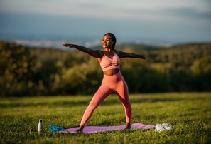 Person in pink workout gear doing yoga pose on a mat outdoors. Green field, trees, and clear sky in background. Water bottle nearby.