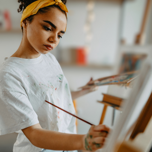 A person with a yellow headband paints on a canvas in a cozy room. A guitar is visible in the blurred background, creating a creative vibe.