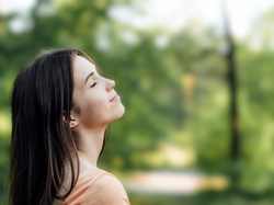 Woman in orange shirt with eyes closed, smiling peacefully in a green, blurred outdoor setting, suggesting a calm and serene mood.