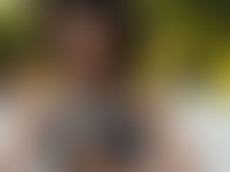 Woman meditating outdoors with eyes closed, wearing a dark tank top. Background is blurred with green foliage, creating a serene mood.