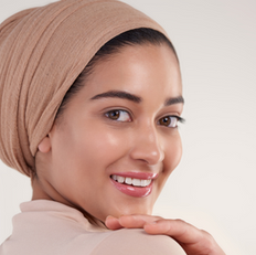 Smiling person in a beige headscarf and top against a soft white background, displaying a joyful expression with a soft focus.