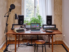 Laptop on wooden desk with audio equipment, typewriter, and plants. Microphone and speakers set for recording. Bright window, relaxed vibe.