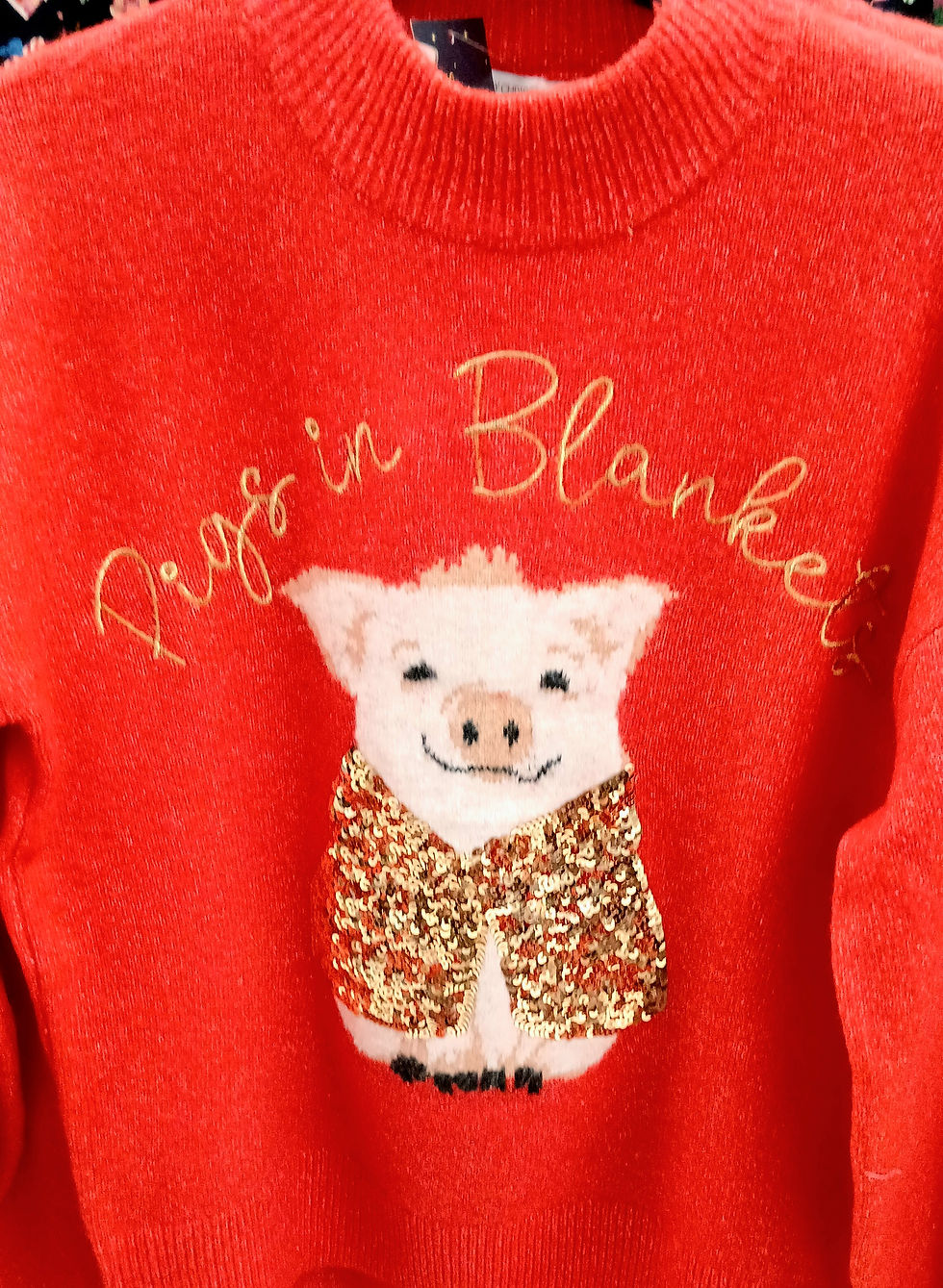 Red sweater with a smiling pig wearing a sequined cloak. Text reads "Pigs in Blankets" in gold. Whimsical and festive.
