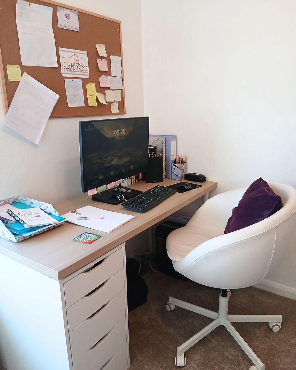 Lilacc Mills' home office with a desk, computer, and swivel chair. A corkboard with notes hangs above. Notebooks and pens are on the light wood desk.